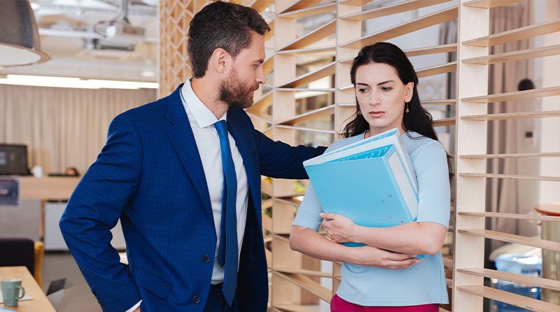 slick young businessman leaning very closely next to an uncomfortable looking female colleague holding stacks of files